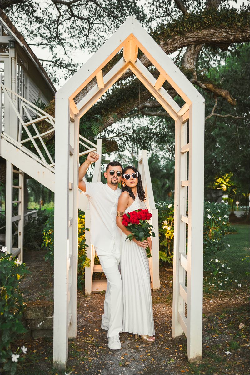 Modern white geometric wedding arch frames couple in matching white outfits against garden setting.