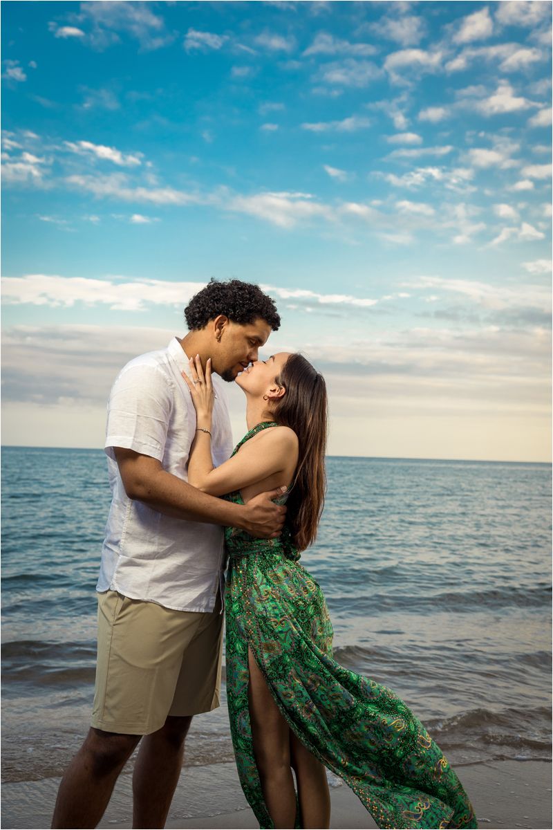 Romantic couple sharing intimate moments by the ocean at sunset with a green floral dress and casual attire.