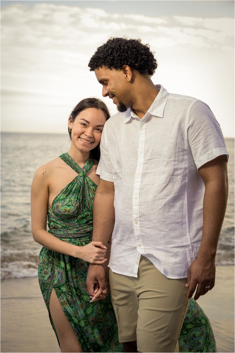 A couple shares a romantic moment on the beach at sunset, wearing a green halter dress and white button-down shirt.