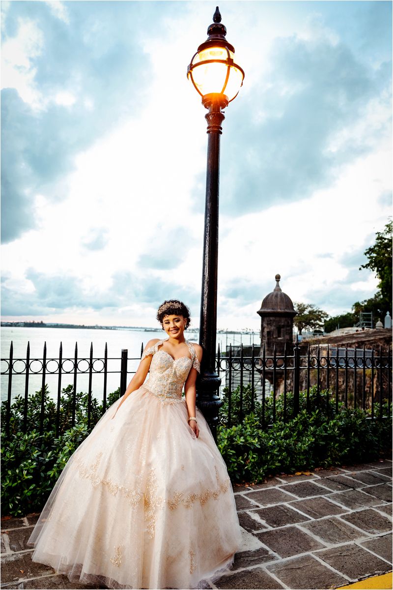 An elegant ball gown with tulle skirt photographed outdoors against a cloudy sky and vintage lampposts.