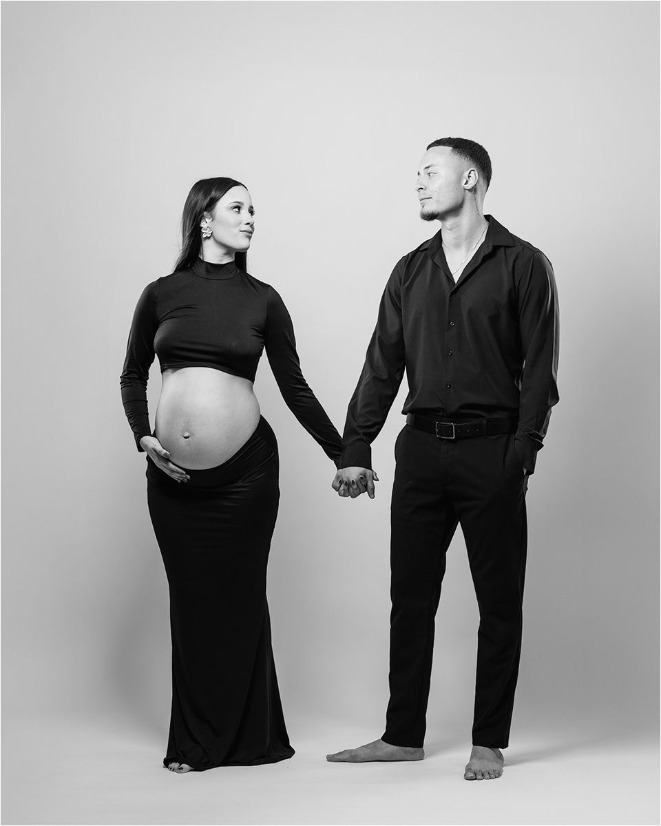 Black and white maternity portrait of a couple in matching dark outfits holding hands against a light backdrop.