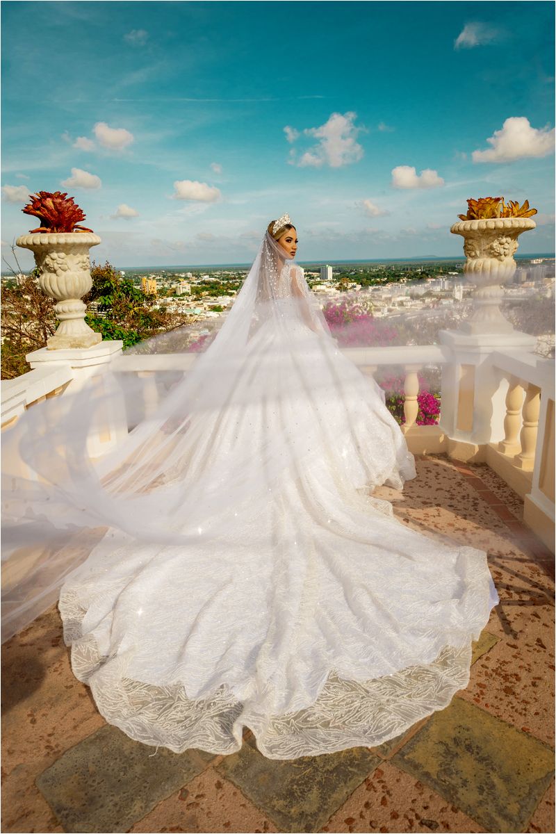 A flowing white lace wedding dress with a dramatic cathedral train displayed on a scenic balcony overlooking a coastal view.