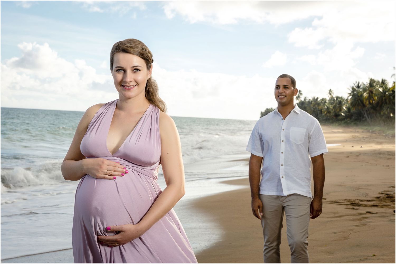 Beach maternity photoshoot with couple standing by ocean waves.