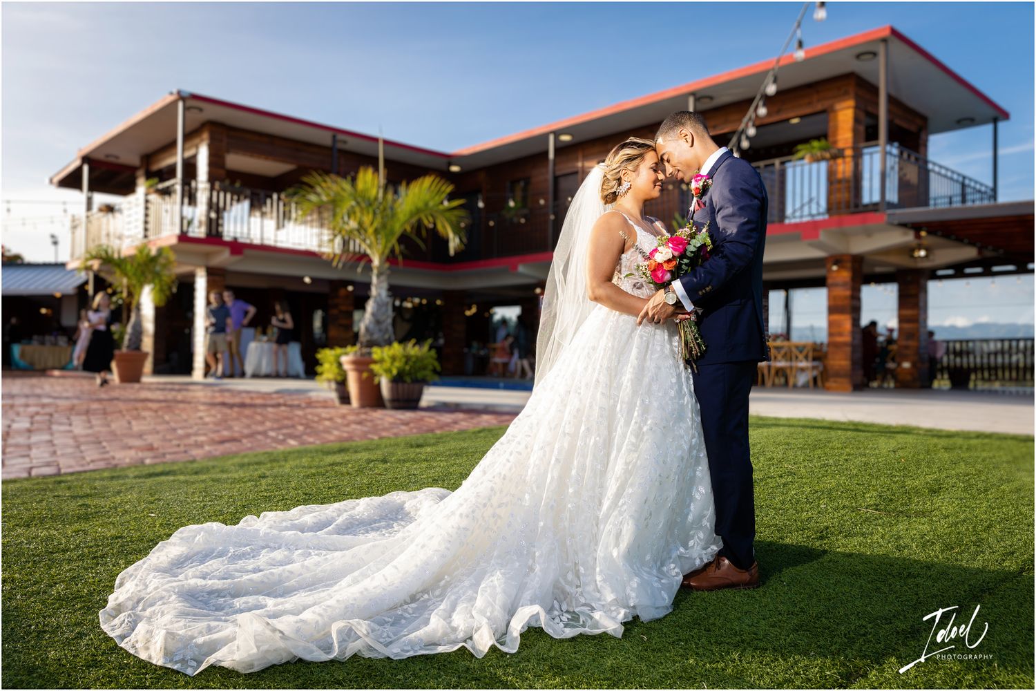 A wedding couple embraces on a manicured lawn in front of a modern two-story venue with palm trees.