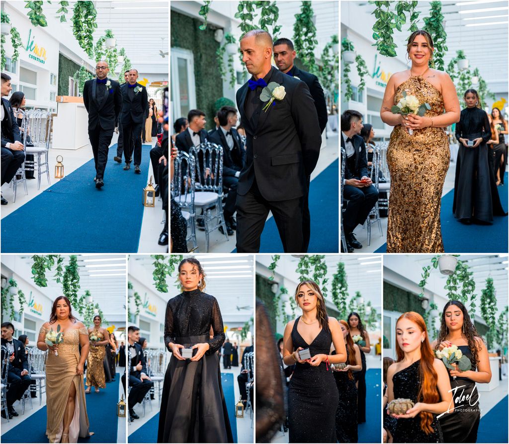 Wedding party members walk down a blue carpet aisle during a ceremony with hanging greenery decorations.