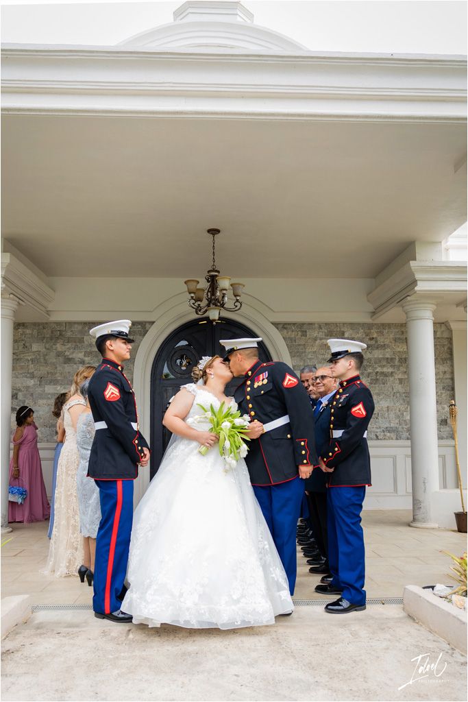 Military service members in dress blues gather around a bride in a white gown outside a columned entrance.