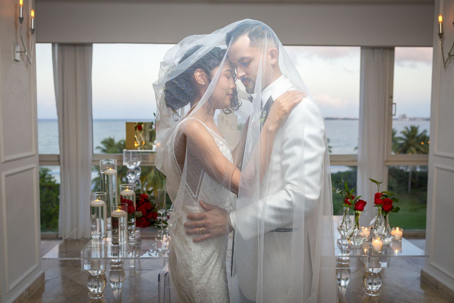 Romantic moment between newlyweds under a veil with ocean view and candlelit decor in background.