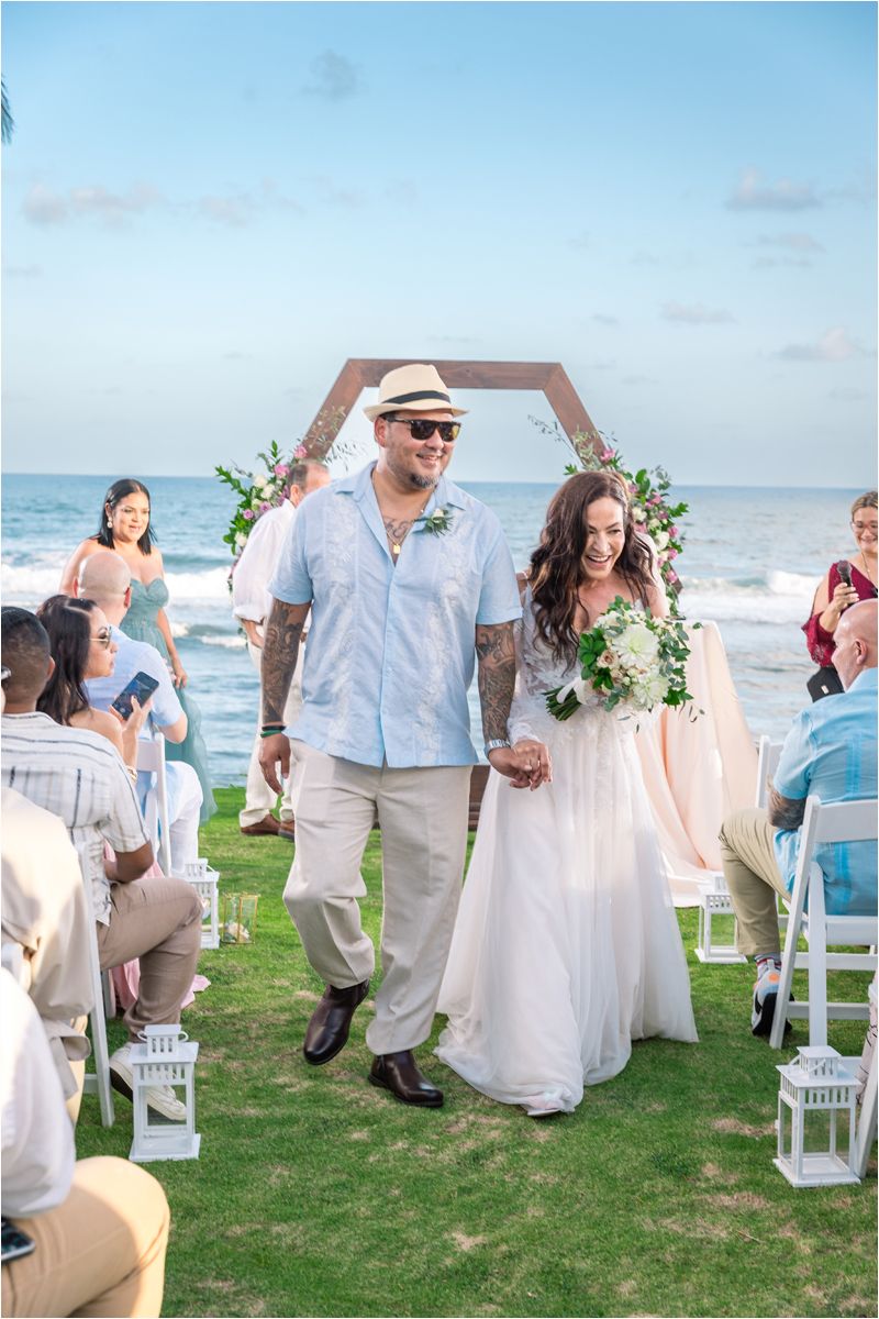 Wedding party dances joyfully on a grassy lawn by the beach with white chairs and ocean views in the background.