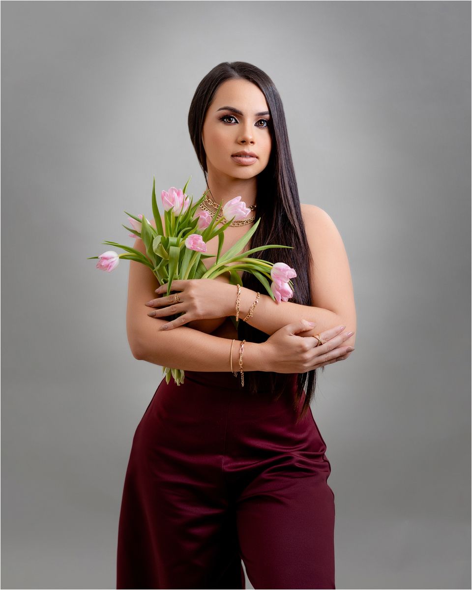 Figure in burgundy dress holds pink tulips against grey backdrop in elegant portrait series.