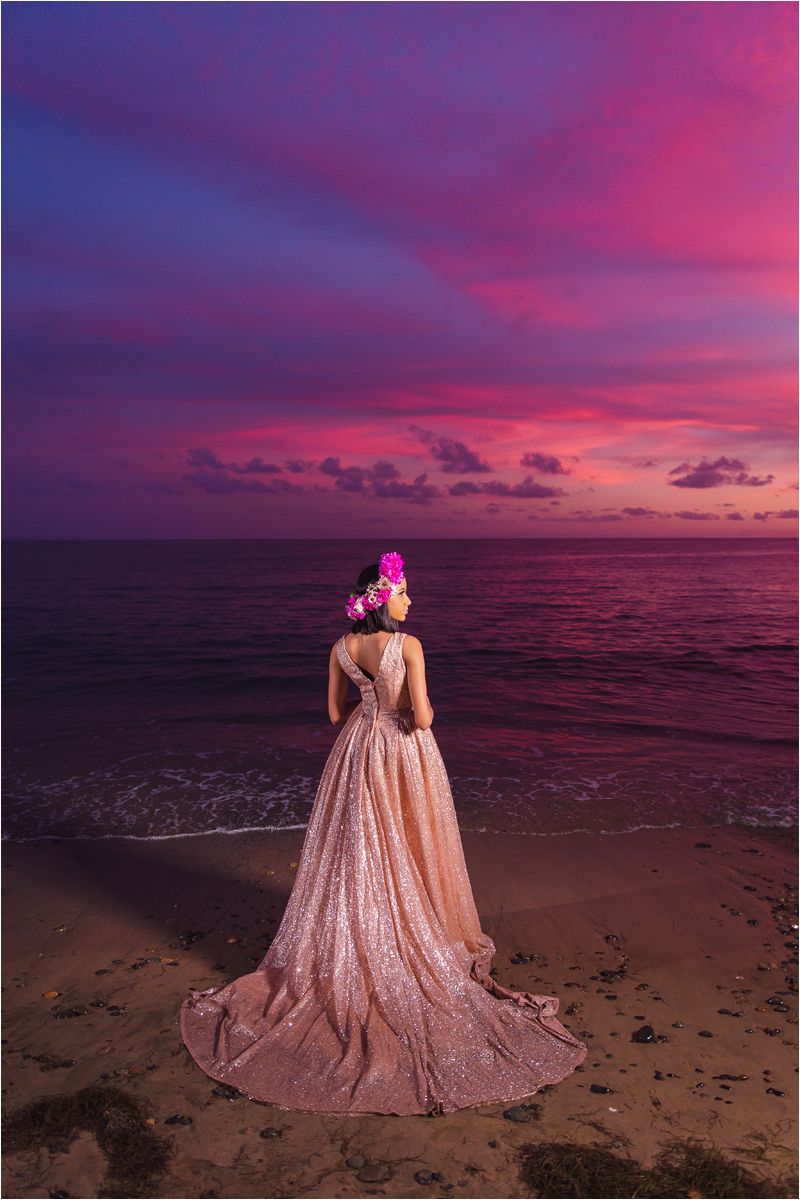 A person in a flowing rose gold gown stands on a beach at sunset with vibrant pink and purple skies.