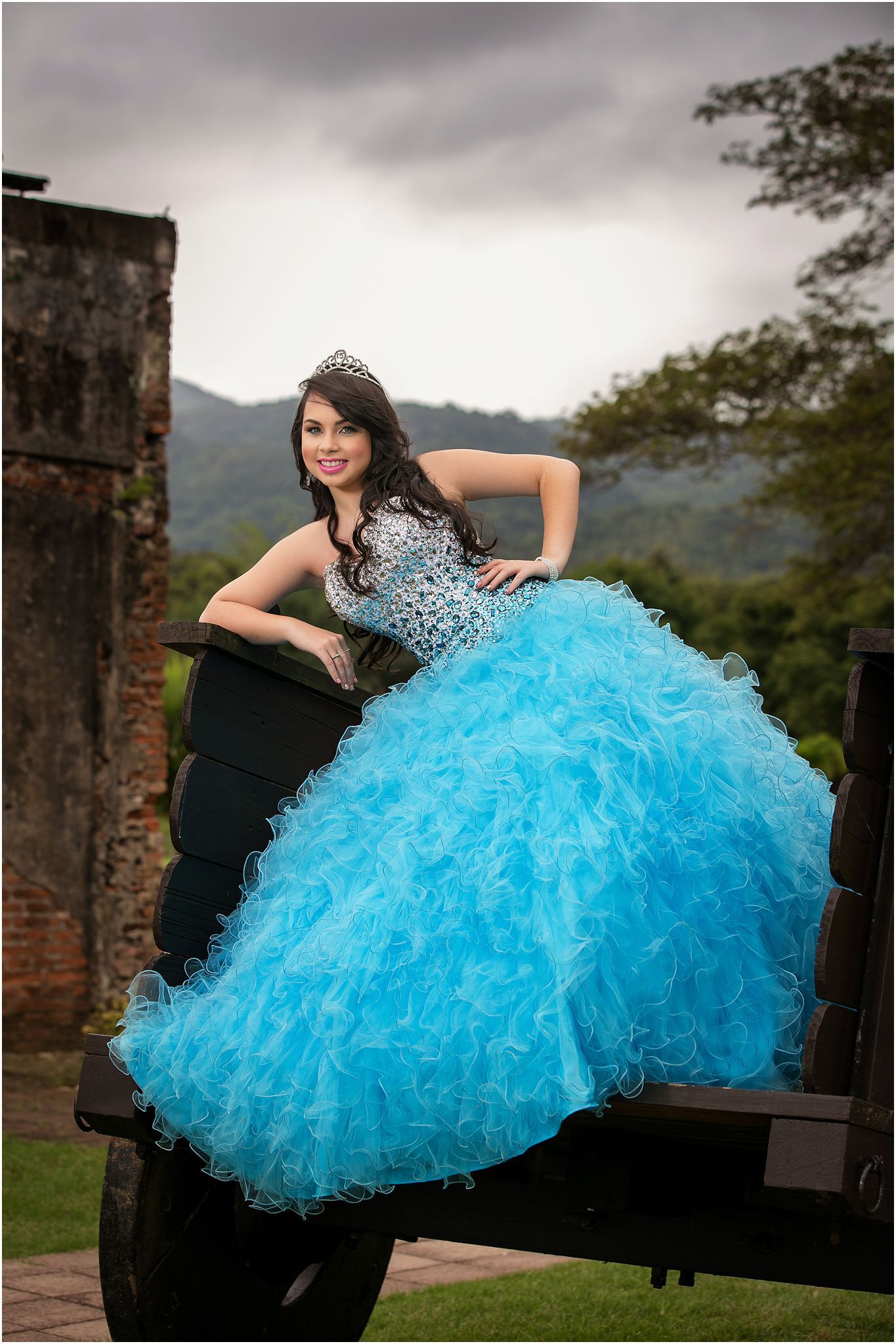 A person in a sparkly turquoise quinceañera dress with ruffled skirt poses on a rustic truck against mountain scenery.