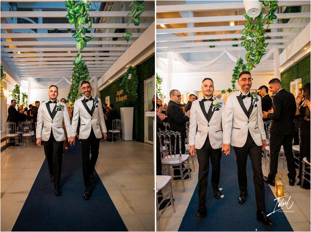 Wedding attendants in white dinner jackets walk down a blue carpet under hanging greenery at an indoor venue.