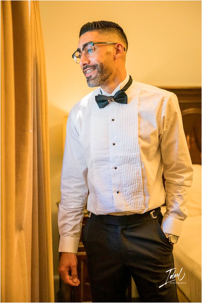 A person wearing a pleated tuxedo shirt, bow tie, and glasses smiles while standing in a warmly lit room.