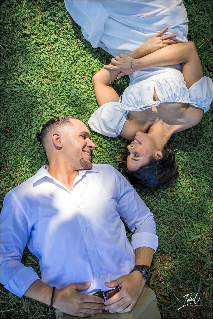 Two people lying on green grass wearing light blue attire share an intimate moment together outdoors on a sunny day.