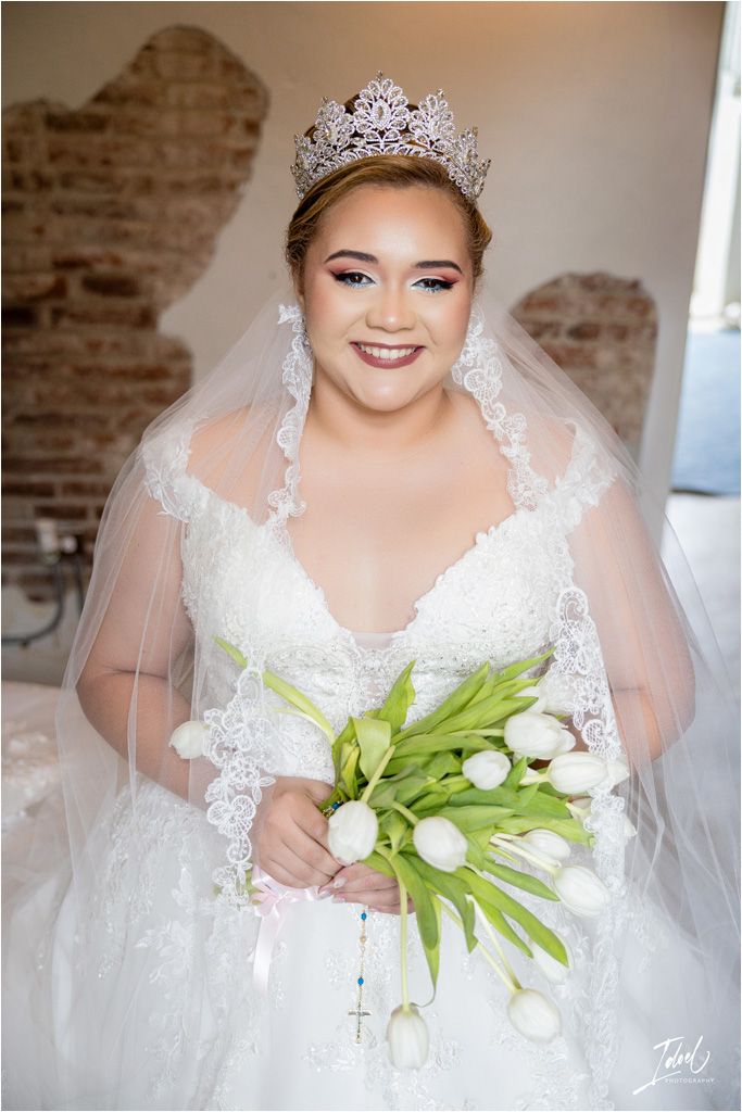 A bride in a white lace wedding gown with a crystal tiara and veil holds a bouquet of white tulips against a rustic brick wall.