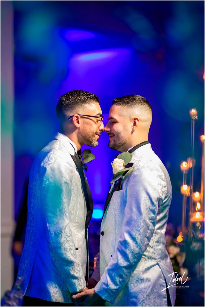 A loving couple shares an intimate moment in matching white jackets under dramatic blue lighting at their wedding celebration.