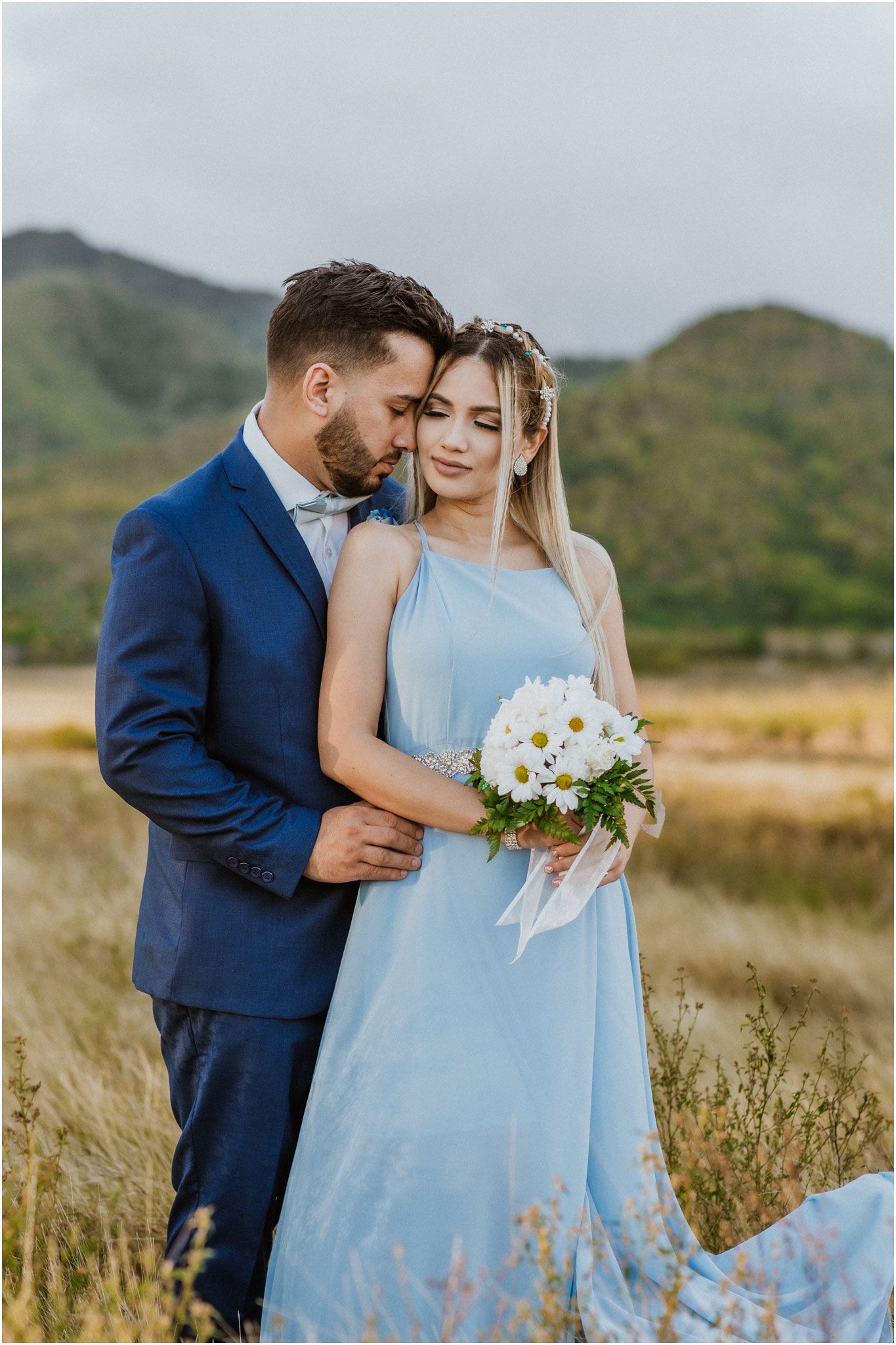 A tender moment between a couple in blue formal wear standing in a grassy field.