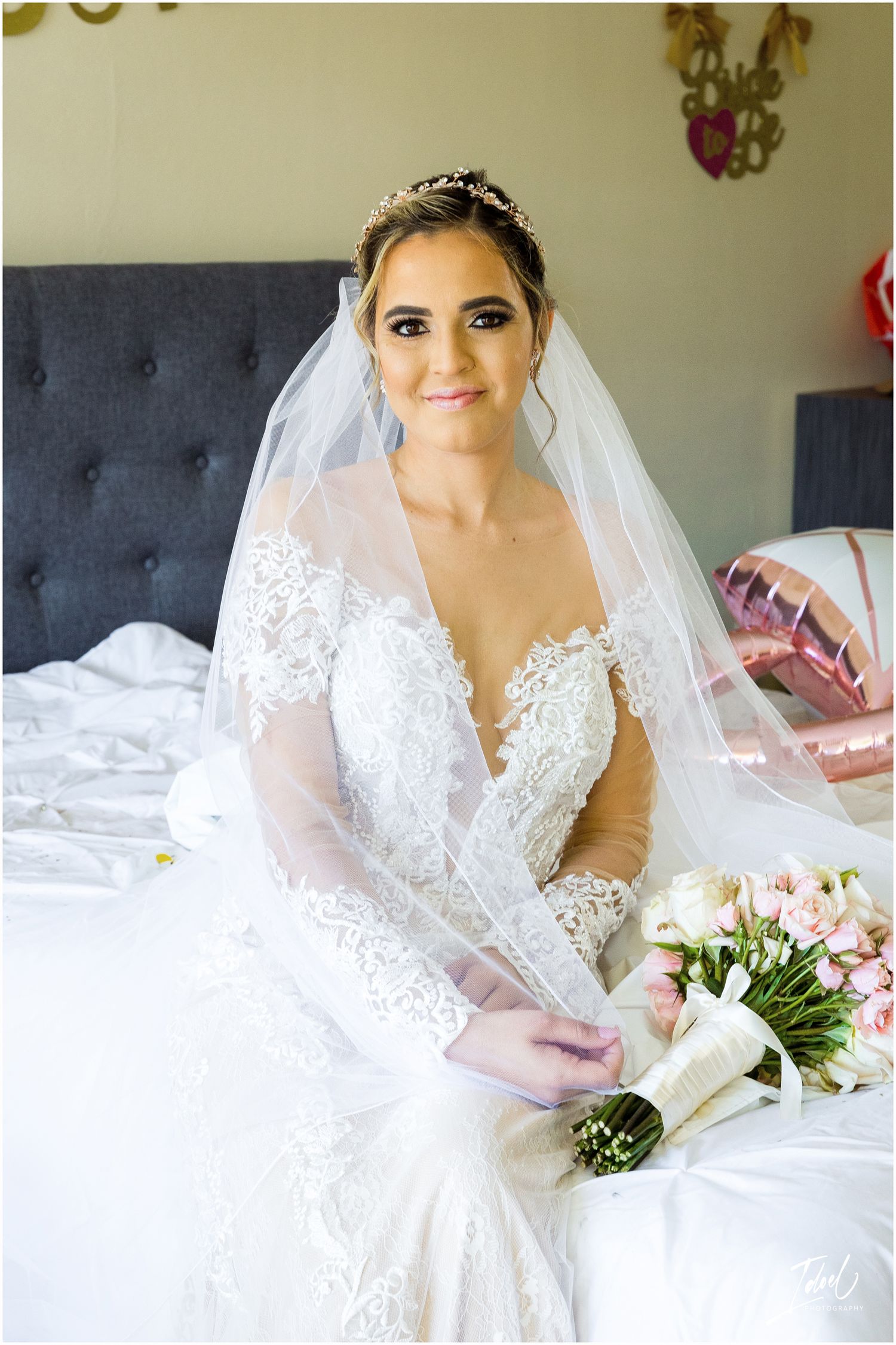 Elegant bride in detailed lace wedding dress poses with floral bouquet against gray headboard.