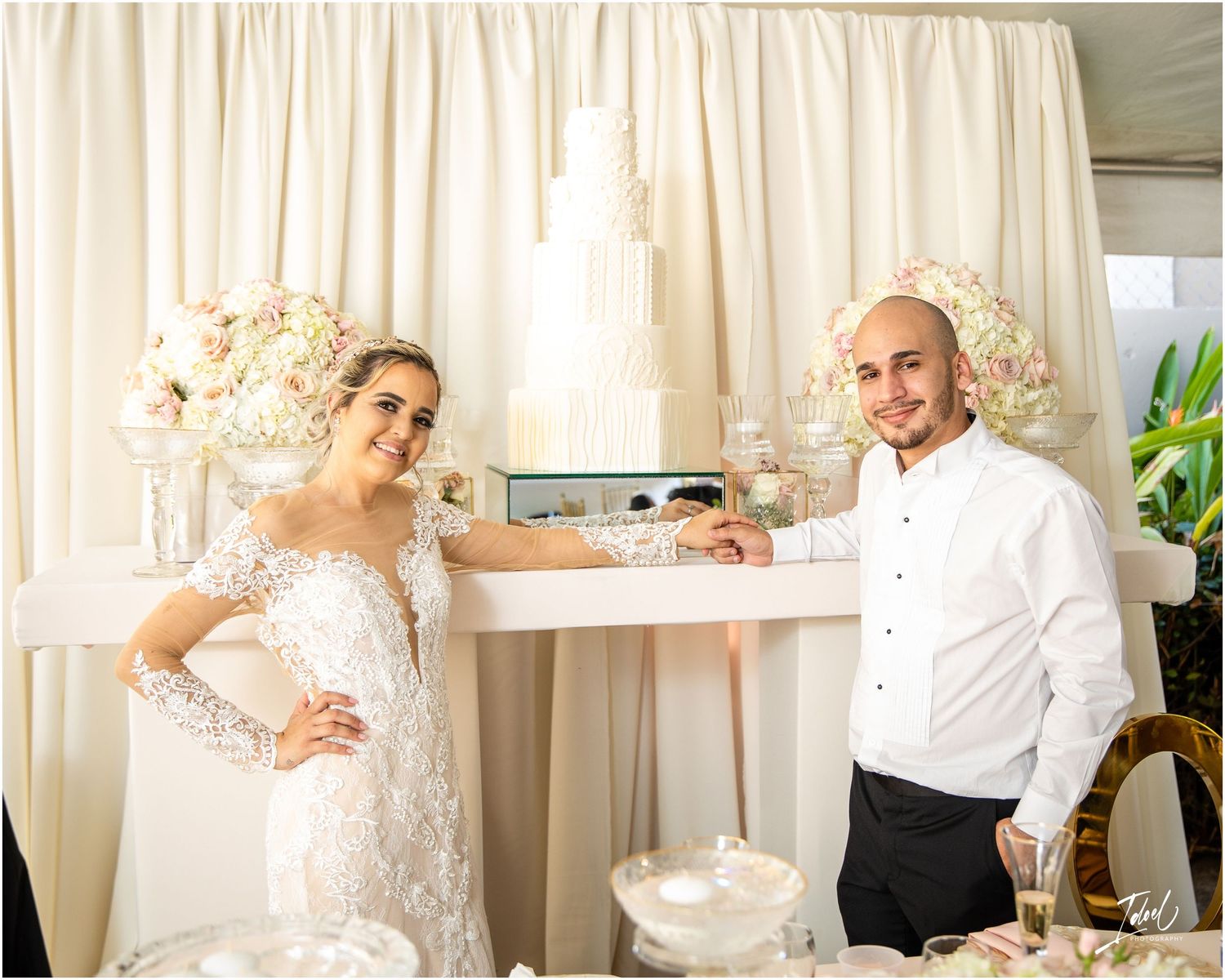 Wedding couple posing together in front of white curtain backdrop with floral decorations.