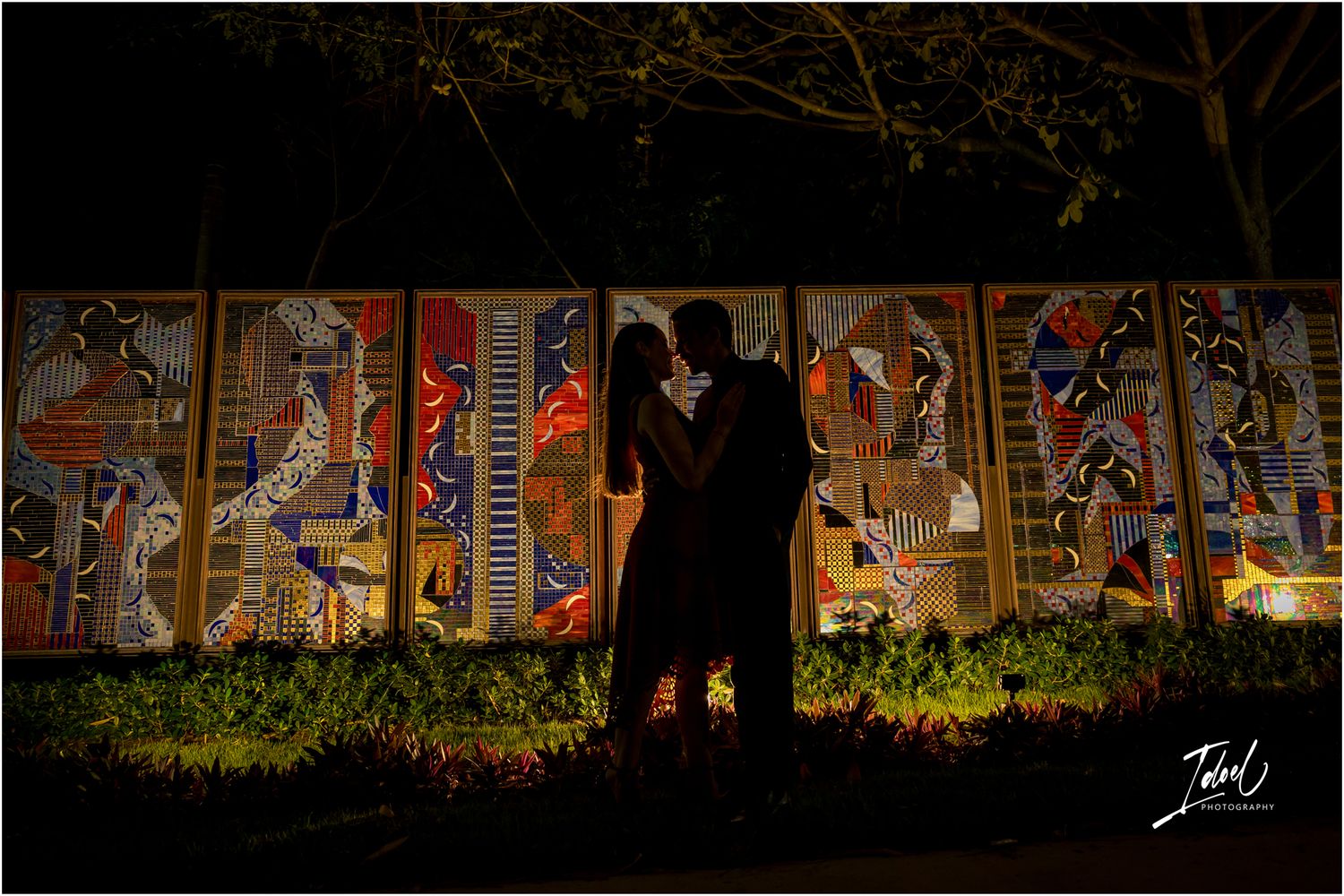 Romantic couple silhouetted at night against vibrant illuminated African mosaic mural artwork outdoors.