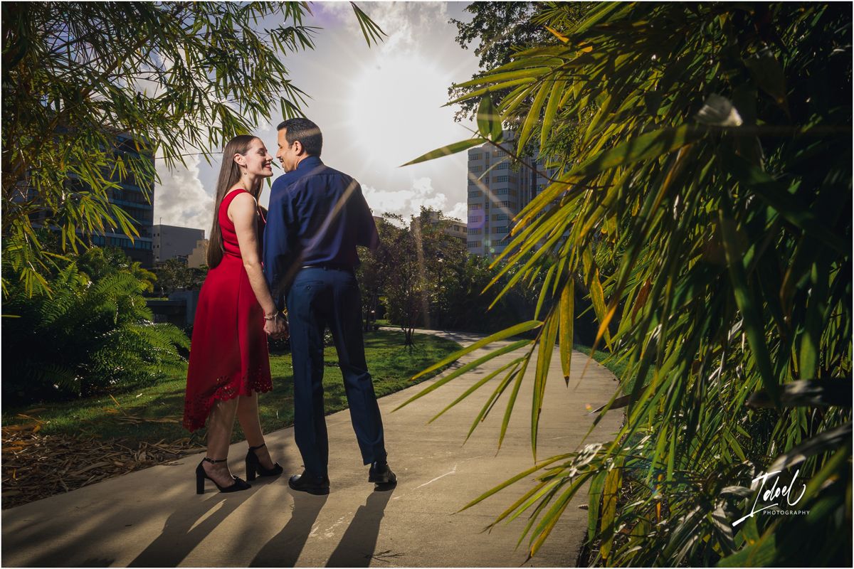 Couple sharing a romantic moment on a garden path, woman in red dress, man in navy suit, with city buildings in background.