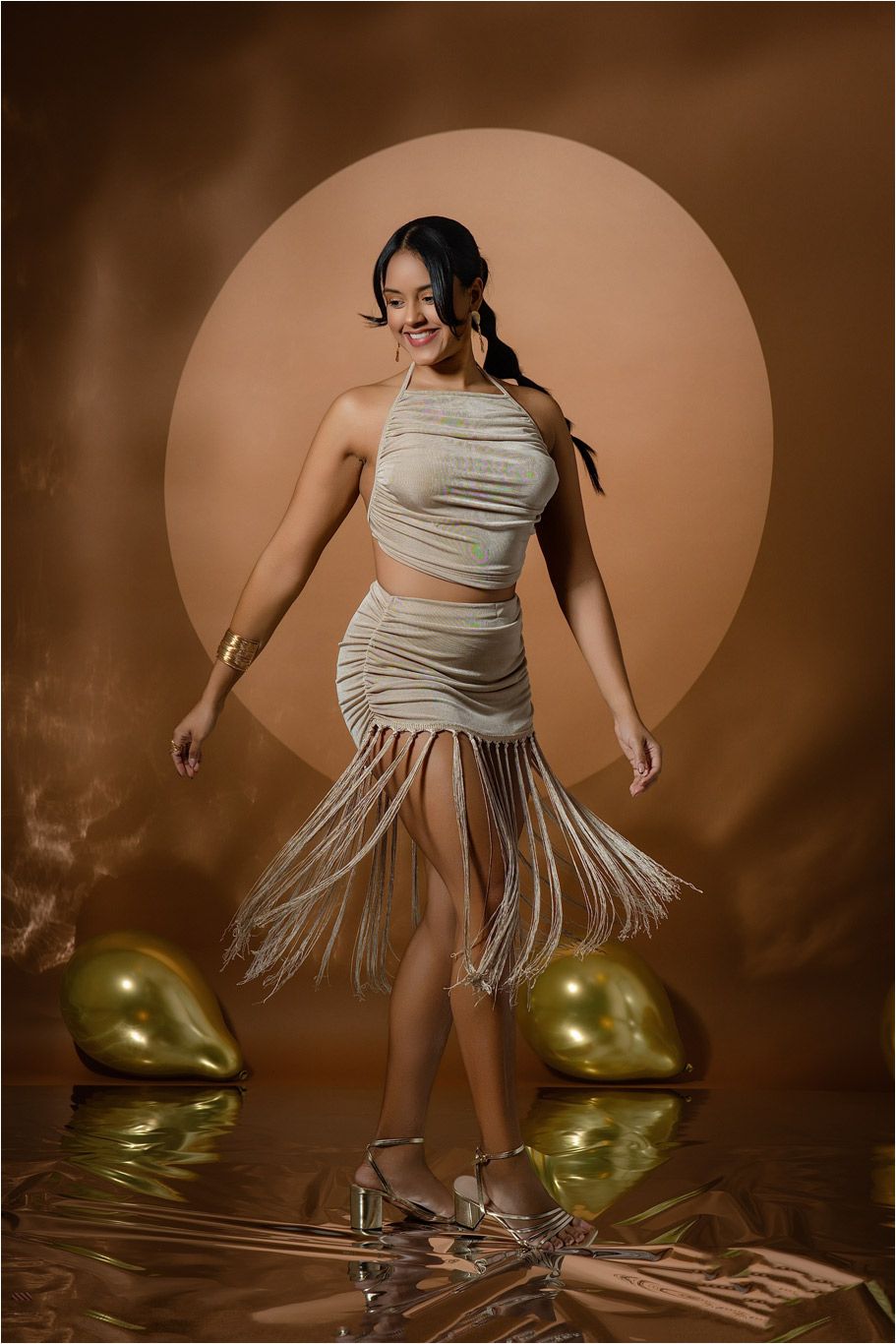 Woman in beige fringe halter crop top and skirt posing with gold balloons on reflective surface against warm brown backdrop.