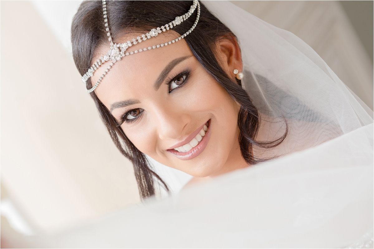 Bride wearing delicate crystal headpiece and flowing white veil smiles warmly in soft natural light.