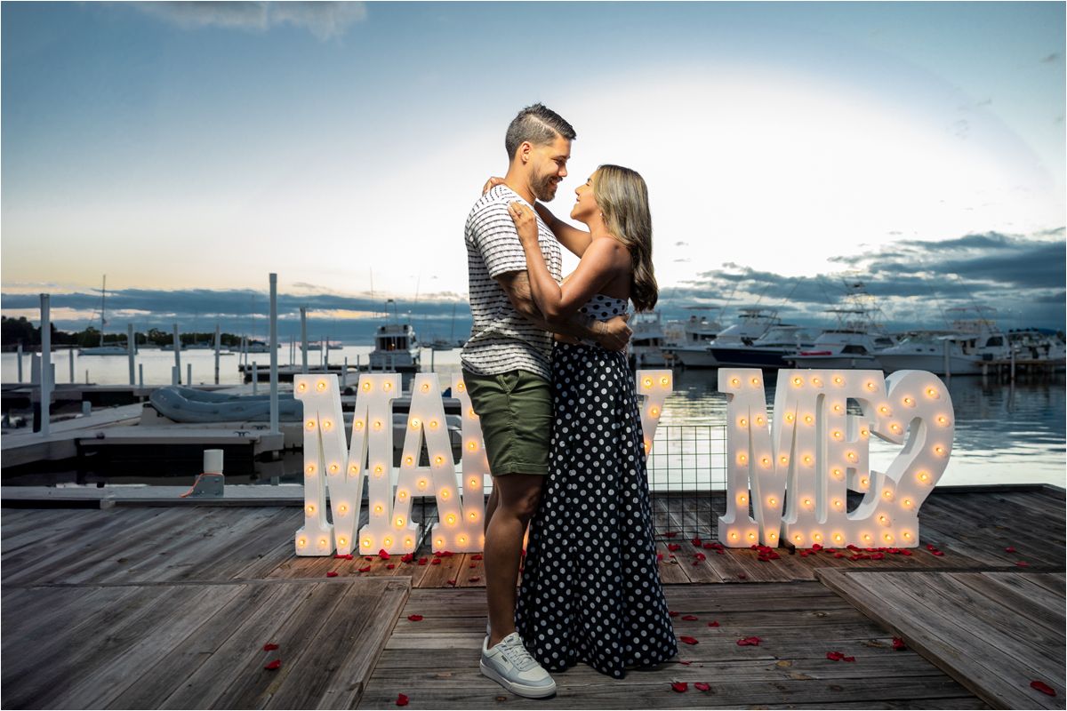 A romantic couple stands on a dock at sunset with illuminated MARRY ME letters creating a marriage proposal scene.