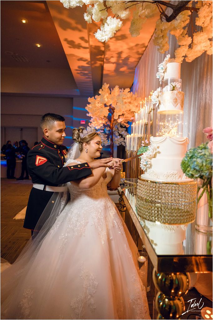 A couple cuts their elegant wedding cake displayed on a crystal stand beneath floral decorations.