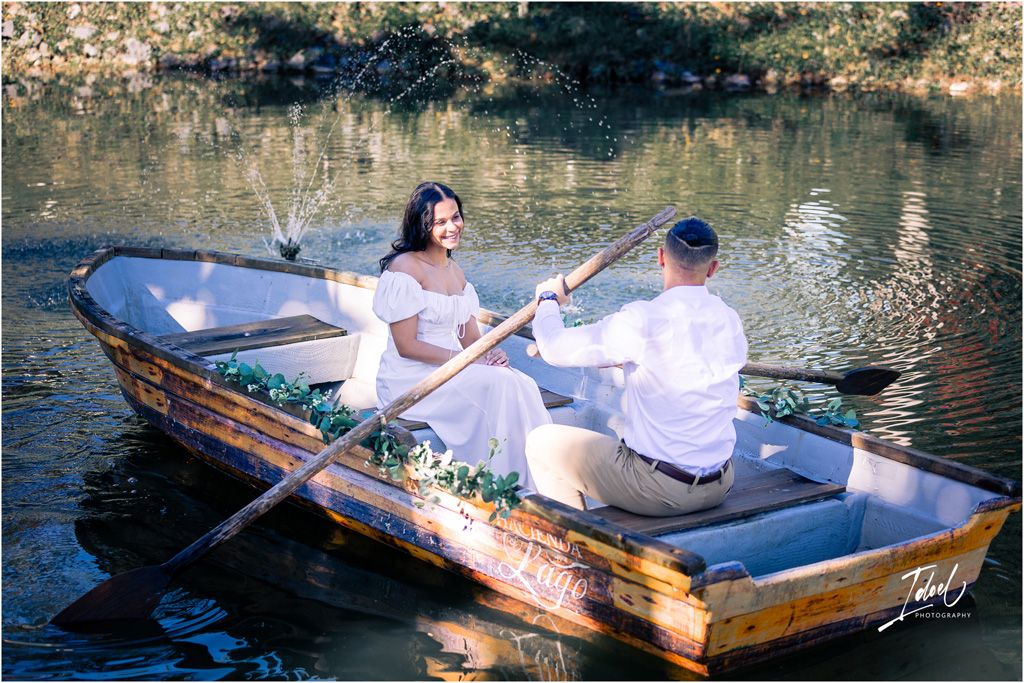 A couple in white outfits share a romantic moment while rowing a wooden boat decorated with flowers on a serene lake.
