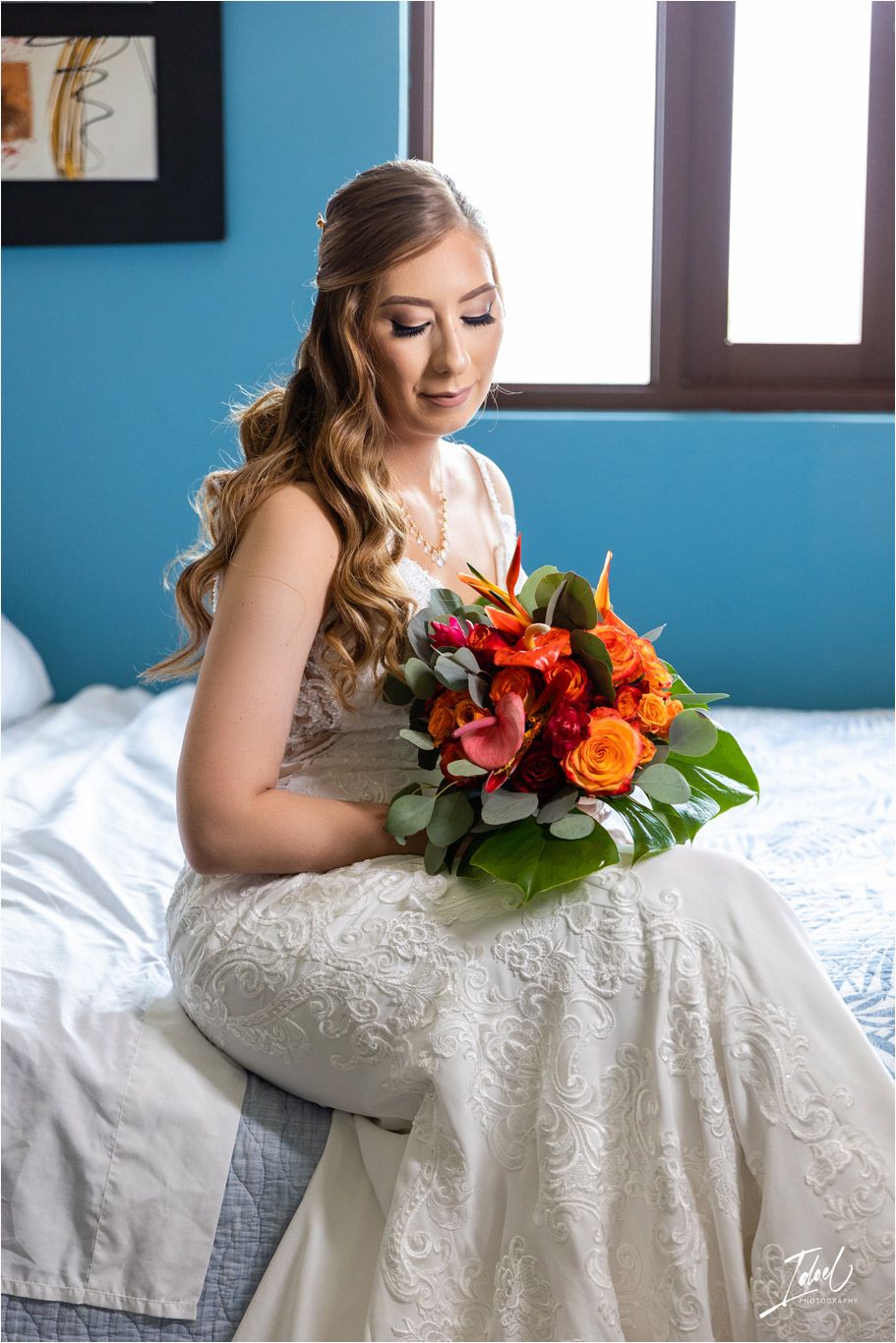 Bride in lace gown holding vibrant orange and red bouquet while sitting on bed.