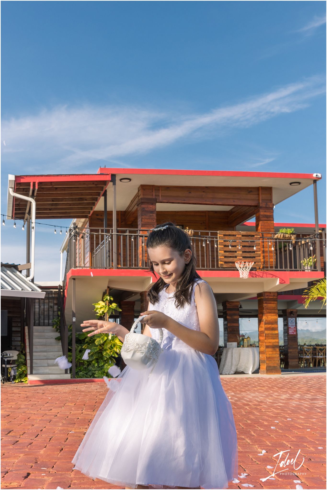 A figure in a white tulle dress checks their phone on a brick patio with a two-story venue in the background.