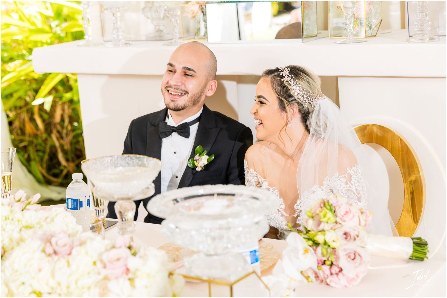 A happy couple laughs together at their wedding reception table surrounded by white floral arrangements.