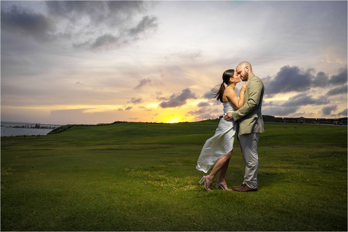 A couple shares a romantic moment in a green field at sunset as a white dress flows in the breeze.