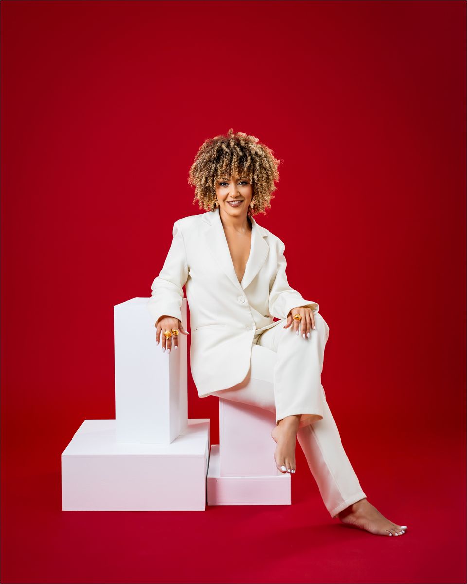 A person in a crisp white pantsuit sits casually on white geometric blocks against a vibrant red background.