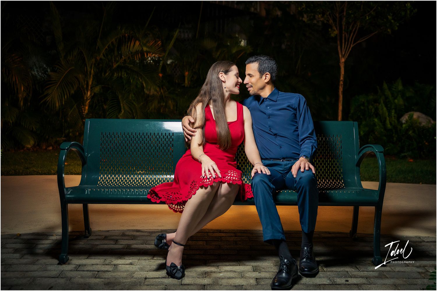 Couple in red dress and blue suit sitting close on a green park bench at night, sharing a romantic moment.
