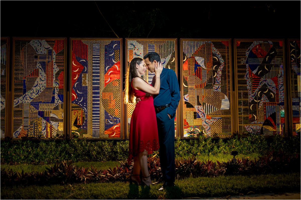 Couple sharing a romantic moment in front of a colorful illuminated mural at night, woman in red dress.