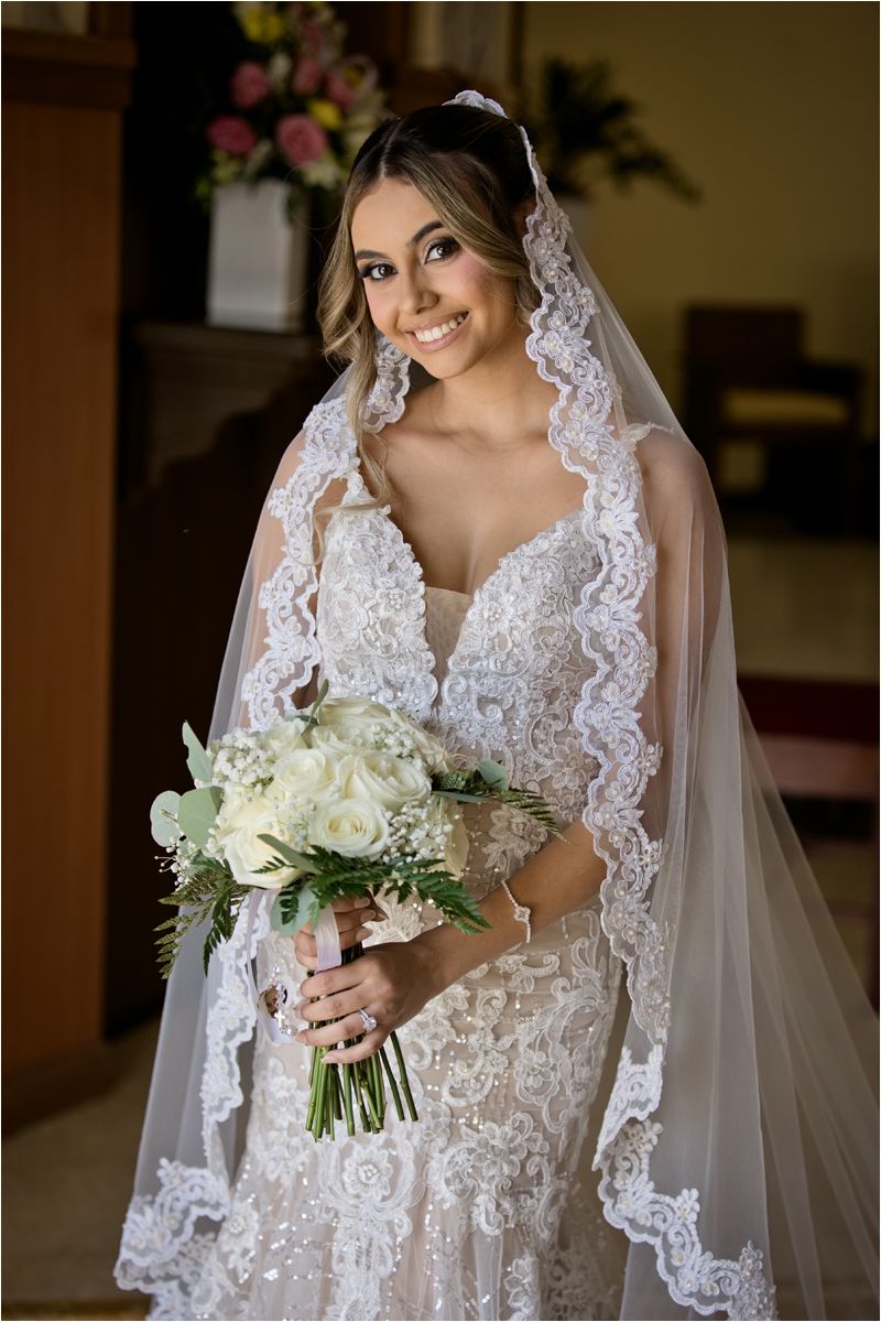 Smiling bride in lace wedding dress and veil holding white floral bouquet inside a church.