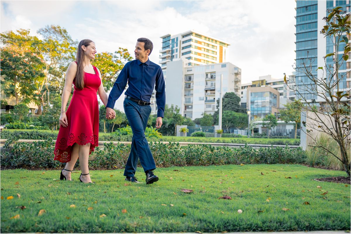 Couple holding hands walking in a lush green park with modern city buildings in the background.