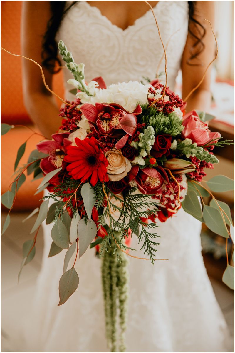 Vibrant red and white wedding bouquet with gerbera daisies, roses, and cascading greenery against a white dress.