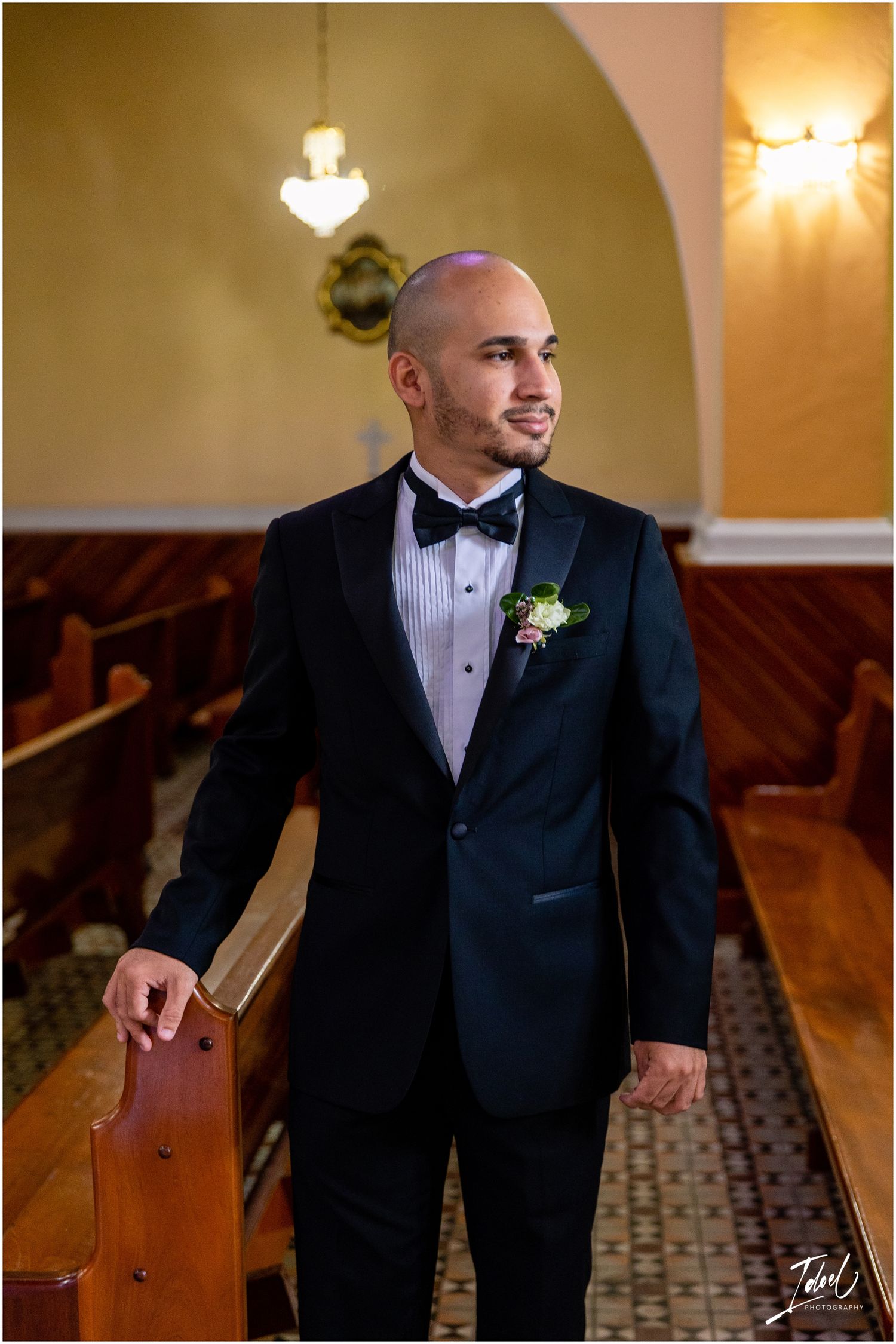 Groom in classic black tuxedo and bow tie stands in church aisle with vintage wooden pews.