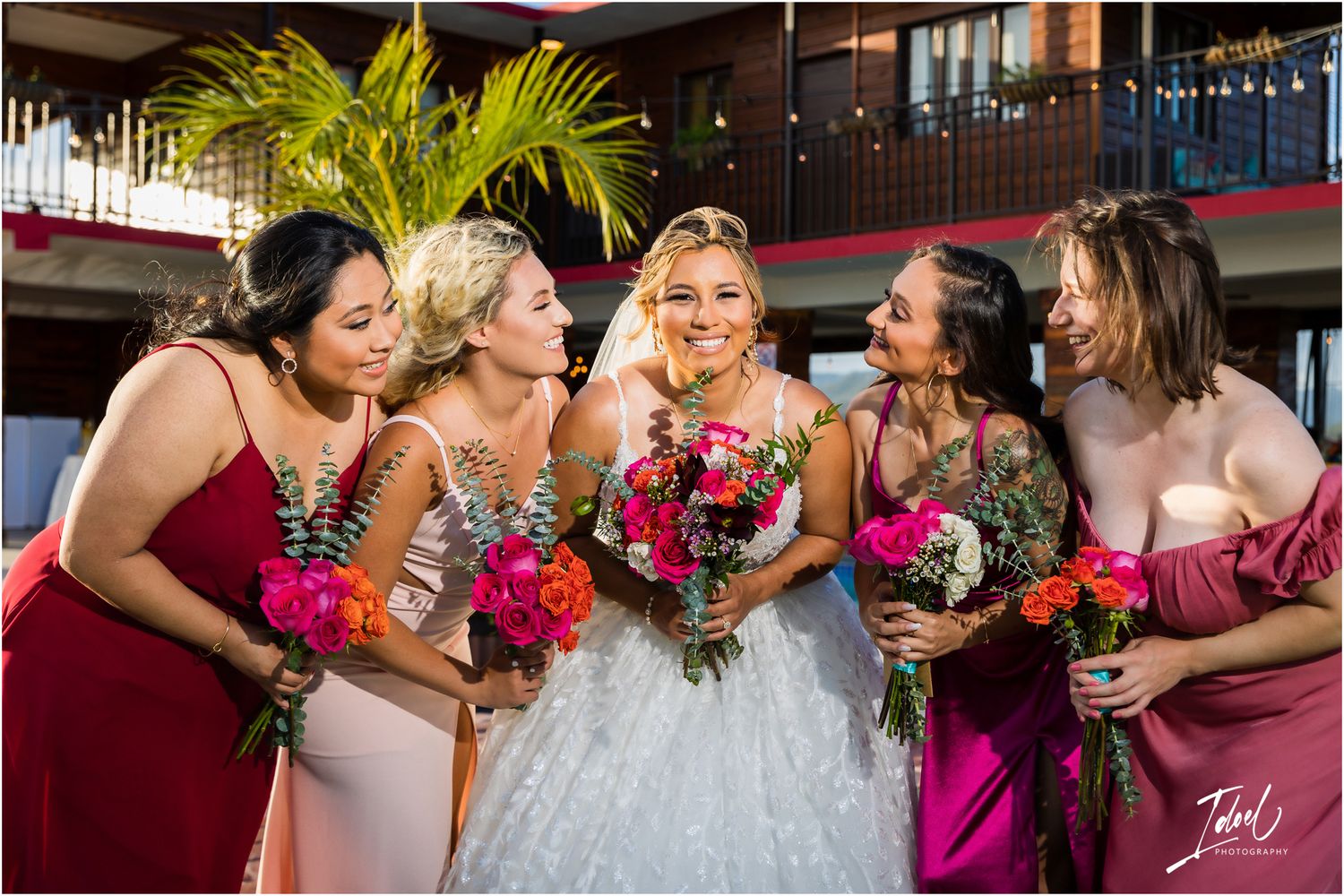 Bridal party shares candid laughter while holding bright pink floral bouquets against modern venue backdrop.