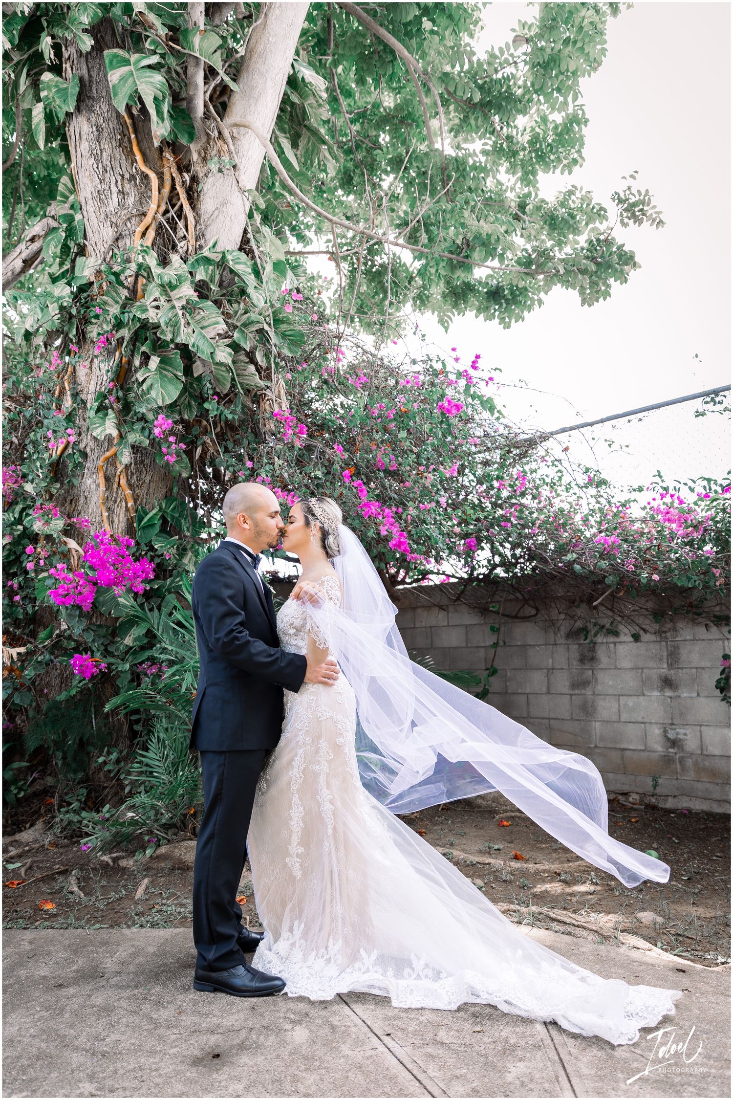 Newlyweds share an intimate moment as the bride's veil flows in the breeze beneath flowering trees.