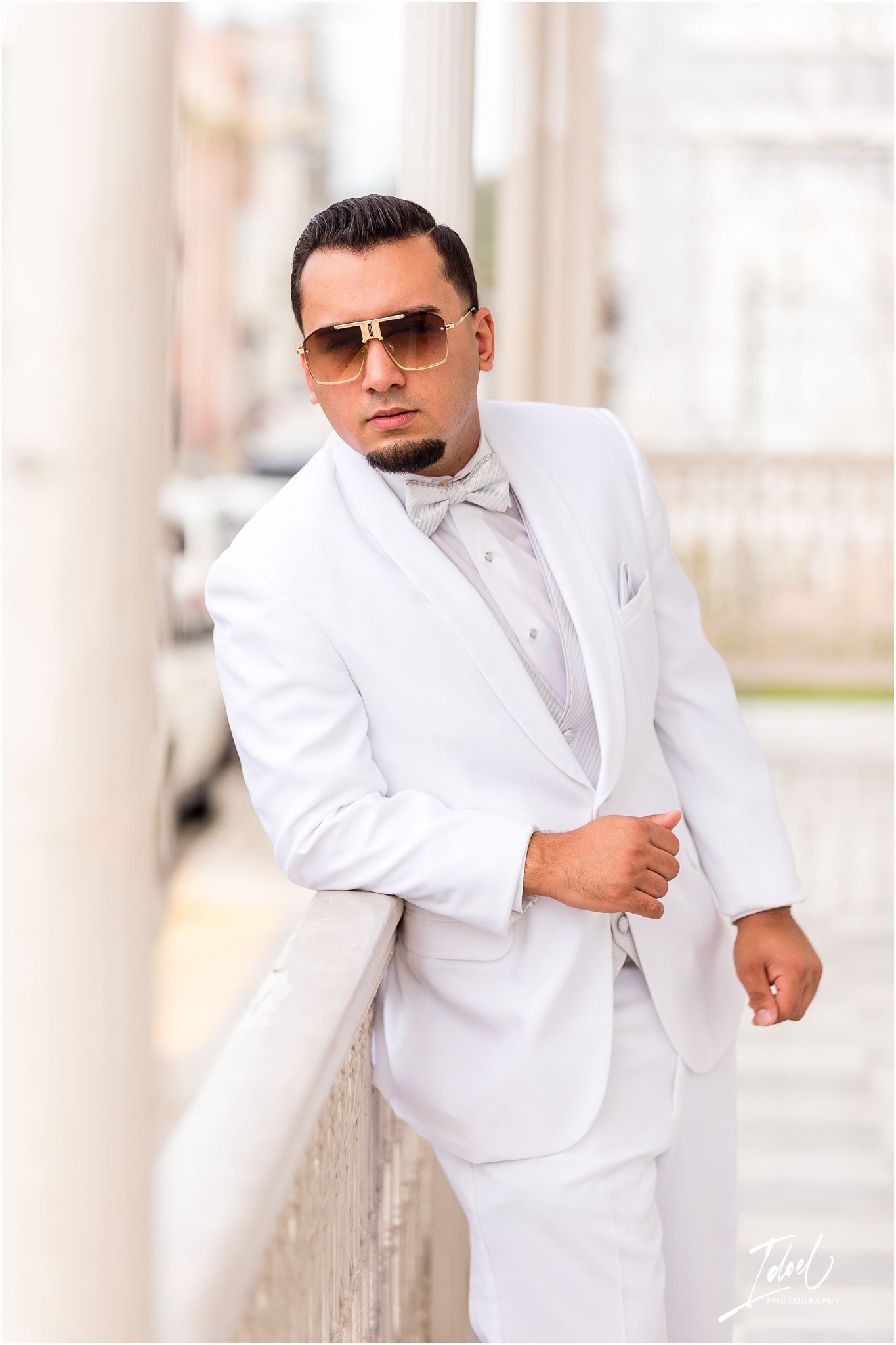Stylish groom in white suit and sunglasses poses against classical columns.