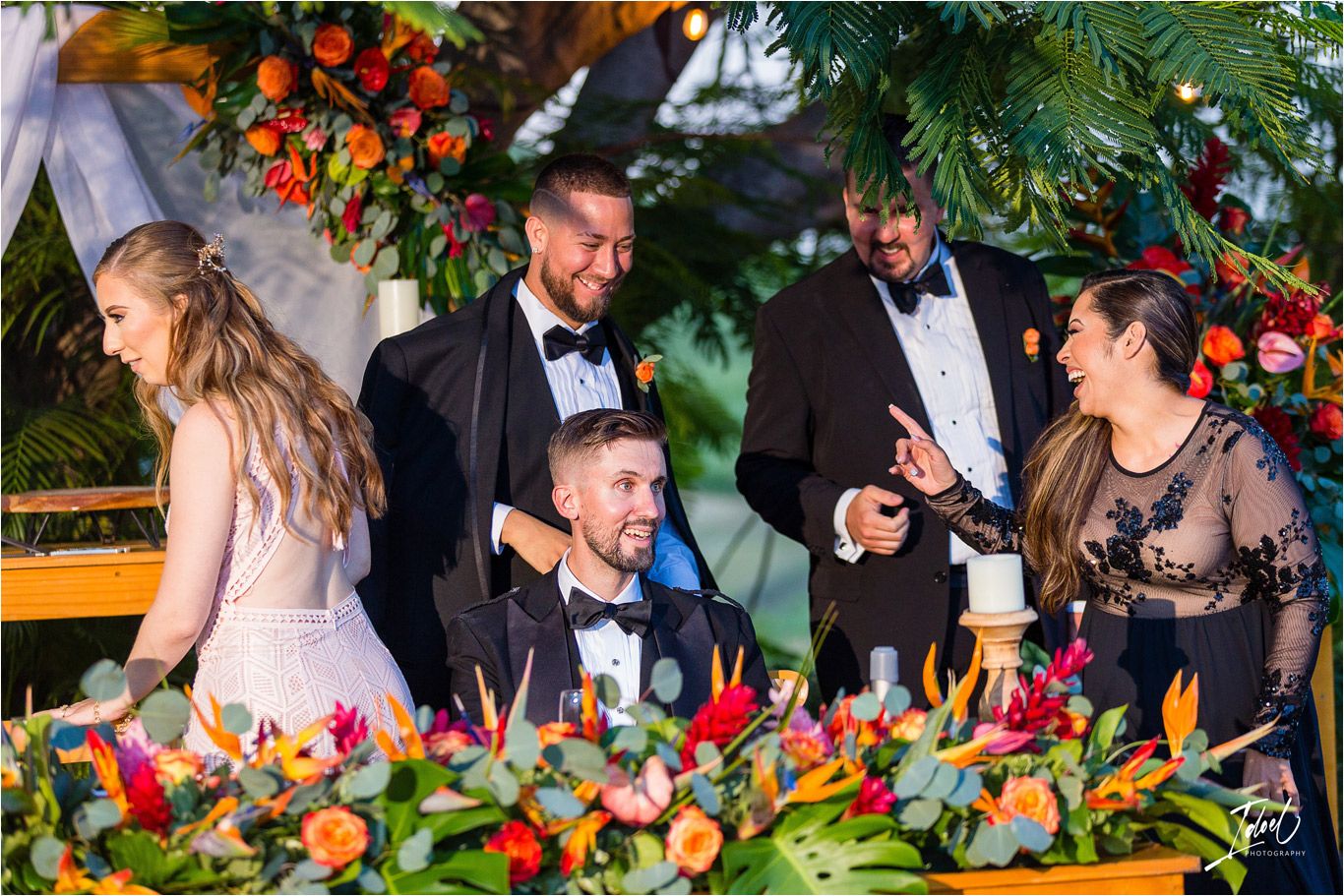 Wedding party celebrates at decorated head table with bright floral arrangements.