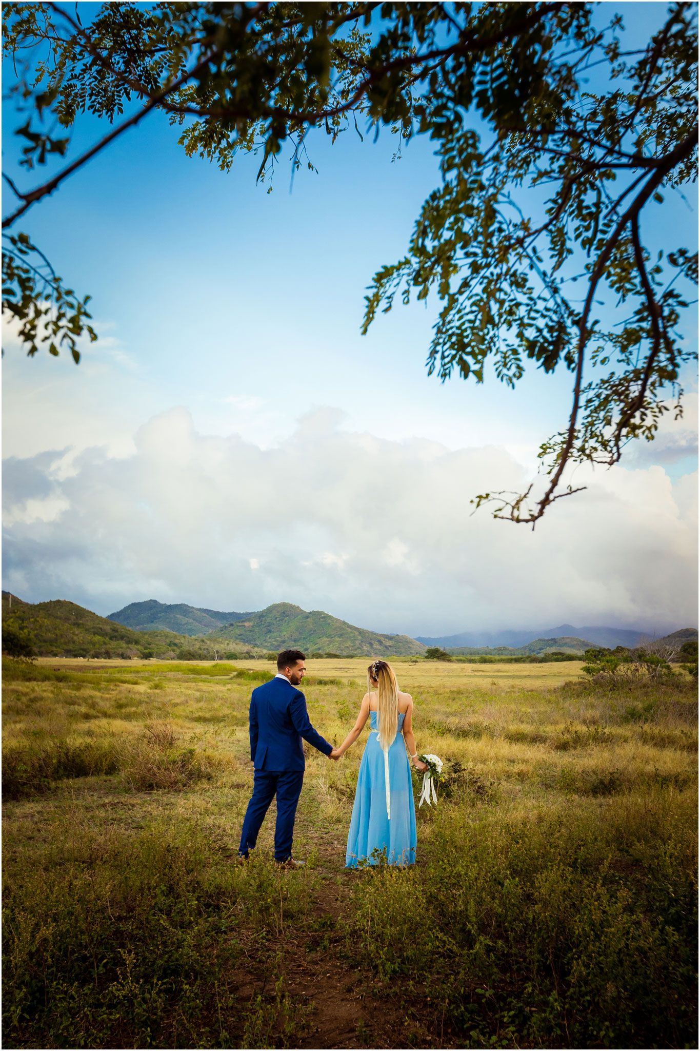 Two people hold hands while standing in a golden meadow with mountains and dramatic clouds in the distance.
