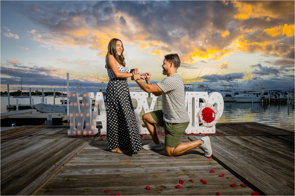 A romantic proposal scene on a marina dock at sunset with rose petals and a decorative white bench against cloudy skies.