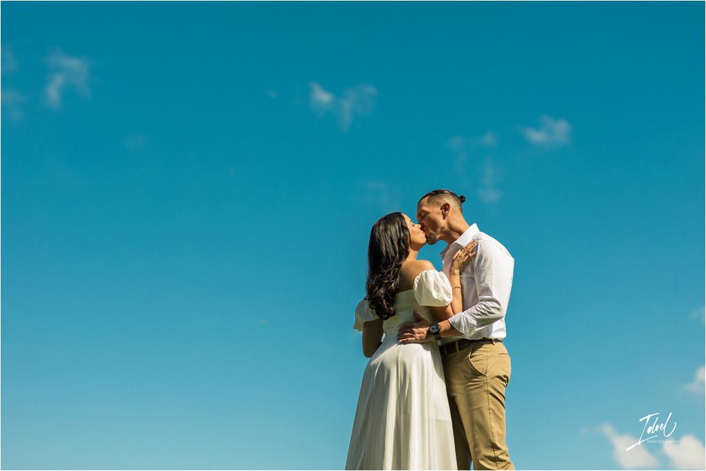 A couple embraces in front of a vibrant turquoise sky with white clouds while sharing a romantic moment.