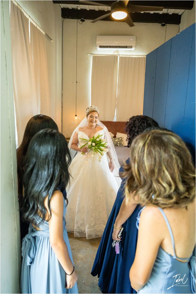 Bridesmaids in blue dresses admire the bride in her wedding gown holding white tulips.