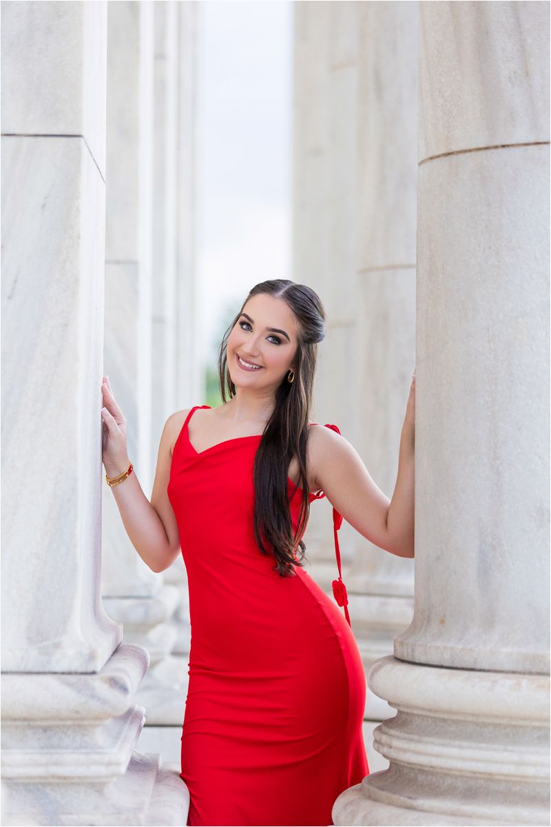 A woman in a red dress poses elegantly between white marble columns in an outdoor architectural setting.